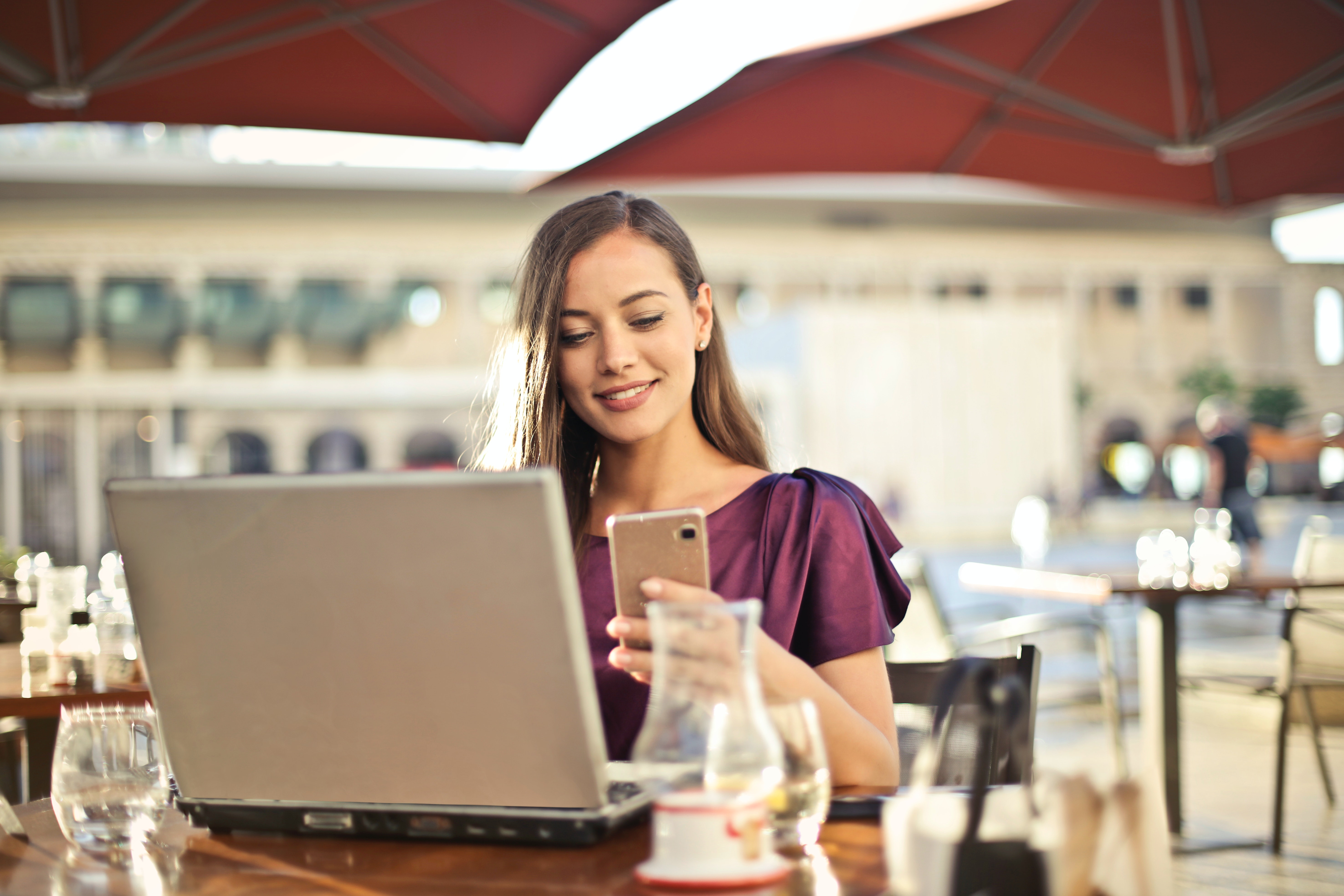 Woman in plum colored top sits outside a cafe on laptop and mobile smart phone