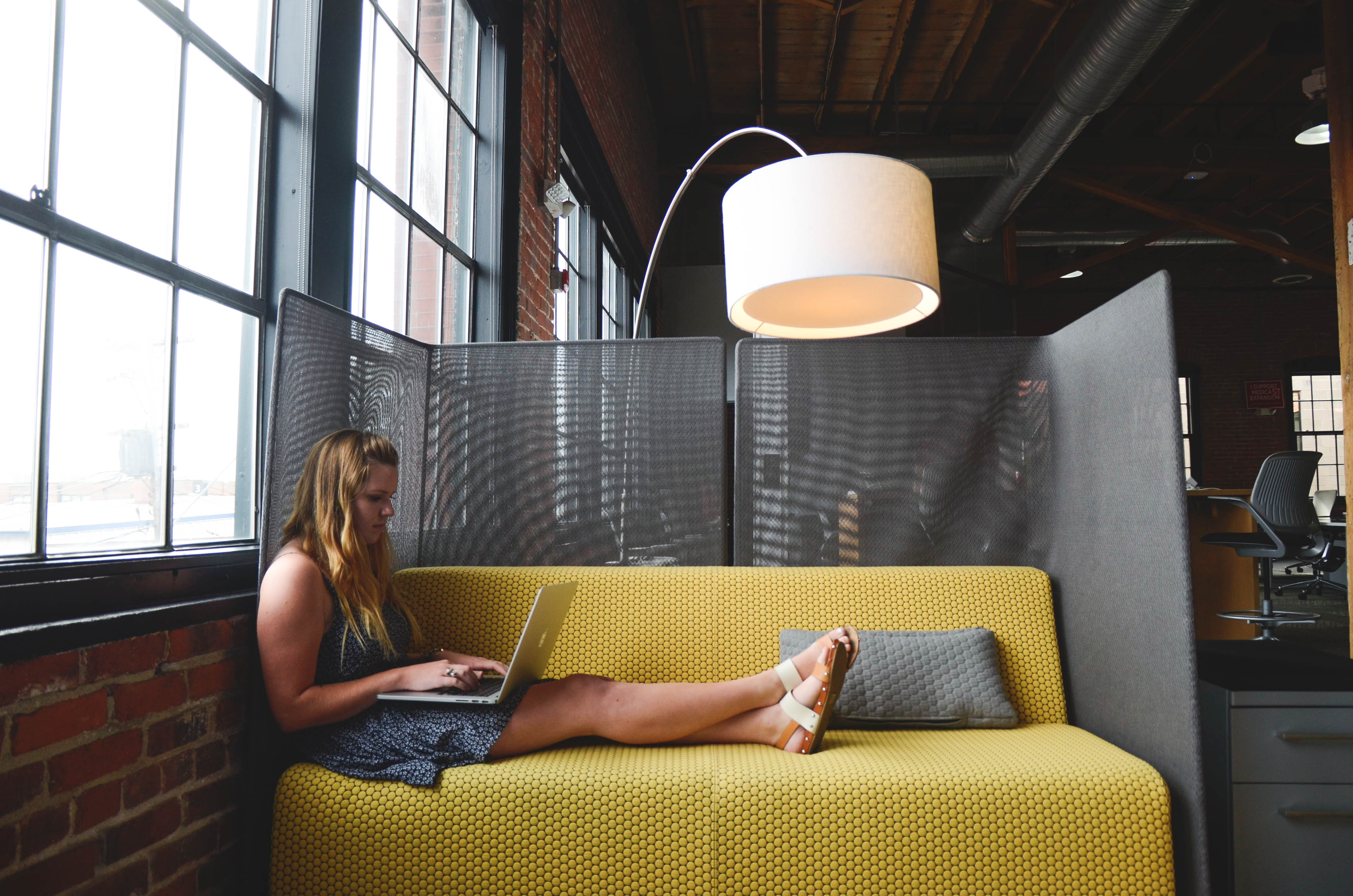 woman sites longways on a yellow couch, working on a laptop