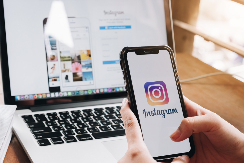 Out of frame person uses mobile smart phone to log onto the Instagram App, while desktop in the background also shows the Instagram login screen.