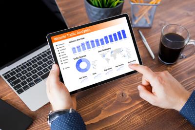 A photograph of a male individual's hands holding a tablet with their left hand, while their right hand points at a data graph and charts displayed on the tablet screen. The screen shows Google Analytics 4 data. The background includes a laptop, a pen, and a pencil placed on a table.