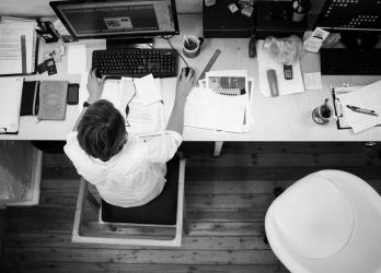 Black & white bird's-eye view of a cluttered desk and overwhelmed worker