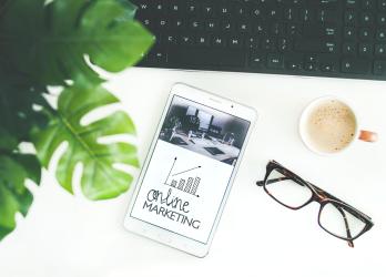 Bird-eye view of desk with plant, coffee, pair of glasses, and phone displaying graphic for Online Marketing