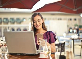 Woman in plum colored top sits outside a cafe on laptop and mobile smart phone
