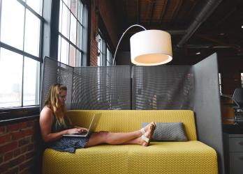 woman sites longways on a yellow couch, working on a laptop