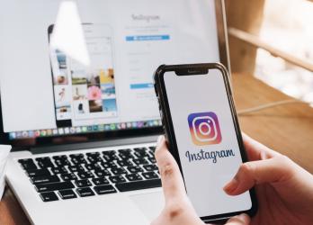 Out of frame person uses mobile smart phone to log onto the Instagram App, while desktop in the background also shows the Instagram login screen.