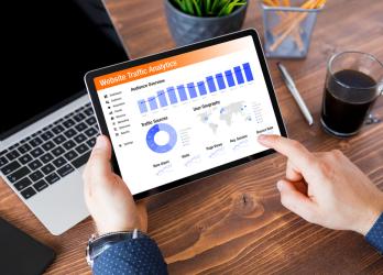 A photograph of a male individual's hands holding a tablet with their left hand, while their right hand points at a data graph and charts displayed on the tablet screen. The screen shows Google Analytics 4 data. The background includes a laptop, a pen, and a pencil placed on a table.